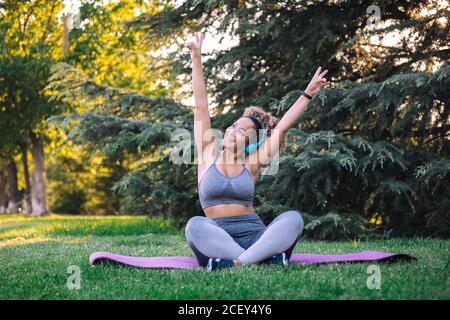 Ottimista giovane etnia femmina con afro hairstyle regolare cuffie mentre ascolto di musica sullo smartphone durante l'allenamento in un parco verde Foto Stock
