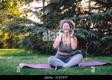 Ottimista giovane etnia femmina con afro hairstyle regolare cuffie mentre ascolto di musica sullo smartphone durante l'allenamento in un parco verde Foto Stock
