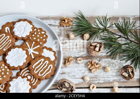 Composizione vista dall'alto con dolci biscotti natalizi fatti in casa decorati con la glassa di zucchero bianco è servita su un piatto posto su un tavolo di legno vicino a varie noci e rami di abete Foto Stock