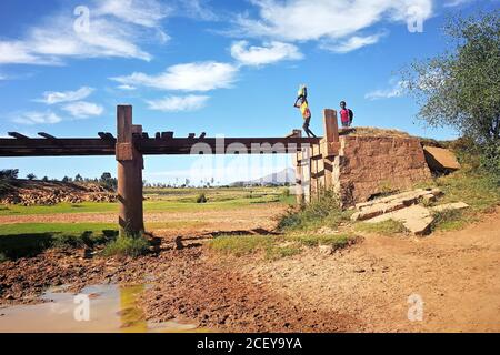 Ambalavao, Madagascar - 27 aprile 2019: Sconosciuto ragazzo malgascio portare borsa sulla testa sopra un ponte semplice, un altro sorridente dietro di lui, tipica Mada Foto Stock