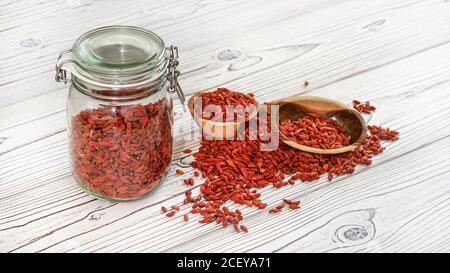 Goji secco aka. Semi di mirtillo all'interno di una bottiglia di vetro, alcuni in ciotole di legno e versato sulla scrivania delle tavole bianche Foto Stock