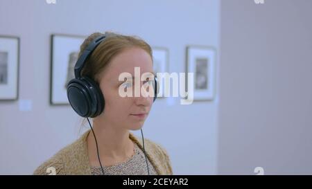 Donna che guarda la esposizione, utilizzando le cuffie e ascolto audio guida in una moderna galleria fotografica. Istruzione e concetto di intrattenimento Foto Stock