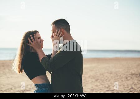 Giovane coppia adulta in piedi sulla costa e spinga i nasi l'un l'altro. Bell'uomo sorridente abbracciando bella donna piena d'amore. Millennial maschile e femminile in posa sulla spiaggia in giornata di sole e guardando via Foto Stock