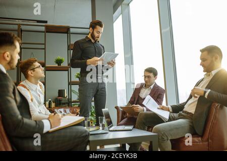 uomo con altoparlante in camicia nera formale che dà voce ai colleghi, tenendo in mano documenti cartacei. isolato in una moderna sala riunioni con panorami Foto Stock