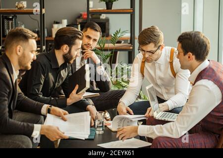 giovani uomini d'affari caucasici in abbigliamento formale che tengono i record sulla carta, facendo appunti e discutere, isolati nella moderna sala riunioni Foto Stock