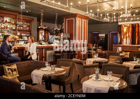 Grande e bellissimo ristorante con tavoli rotondi e sedie morbide, sala illuminata brillantemente da molte piccole lampade, giovani si siede e bevande bianco profumato Foto Stock