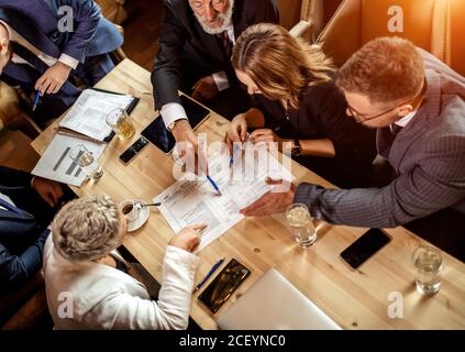 Due belle giovani donne sedute alla scrivania per conferenze di colore chiaro discutono di problemi insieme a uomini d'affari che lavorano sodo in ufficio, via ad alta angolazione Foto Stock