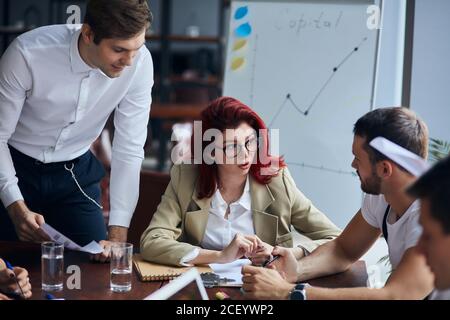 Donna leader fiducioso e attraente che indossa occhiali guardare colleghi e carte, spiegare. Lavoro di squadra in un moderno business center, ufficio Foto Stock