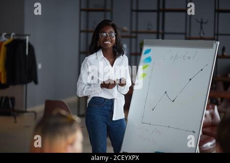 Donna africana personable in occhiali, indossando camicia bianca e jeans, utilizzando diagrammi, motivata afro-americana speaker coach presente business plan Foto Stock