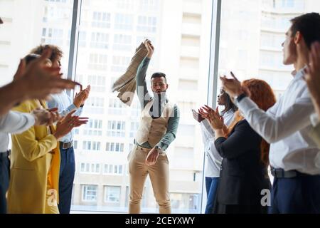 I giovani imprenditori celebrano la vittoria del loro collega che si trova al centro in posa del vincitore con il blazer nelle mani, sfondo della finestra panoramica Foto Stock