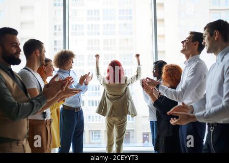 I giovani imprenditori celebrano la vittoria del loro collega in piedi al centro in posa del vincitore con le braccia in su, sfondo della finestra panoramica Foto Stock