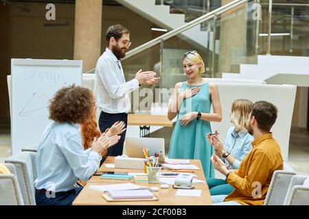 L'uomo alto e non rasato insieme ai lavoratori del team clevel applaudisce giocosamente i giovani donna affascinante con sorriso amichevole in agenzia di stilisti confortevoli Foto Stock