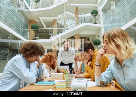 Elegante uomo d'affari nobile con barba spessa e bicchieri rotondi, vestito in elegante camicia bianca si appoggia sul tavolo, parla con i dipendenti intelligenti con ex positivo Foto Stock