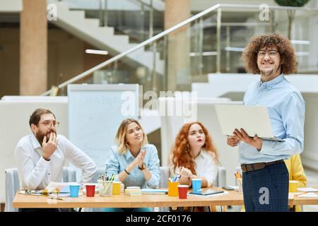 Piacevole Tall uomo non rasato in divertenti occhiali rotondi, espresso piacere, sembra soddisfatto direttamente alla macchina fotografica, mantiene bianco computer portatile, sorride gent Foto Stock