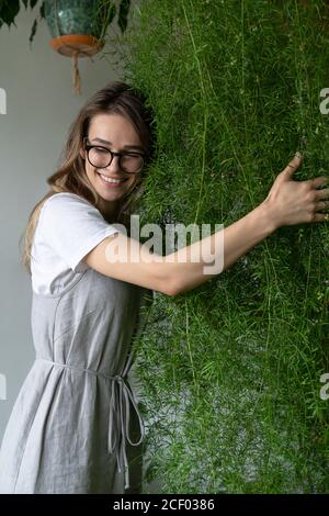 Felice giovane donna giardiniere in abito lieno, abbracciando lussureggiante asparagi felce pianta domestica nel suo negozio di fiori. Verde a casa. Amore delle piante. Interno accogliente Foto Stock