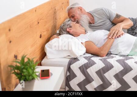 L'uomo anziano dell'hipster bacia la moglie mentre si sdraia a letto - felici persone mature che si divertono a casa - gioiosi anziani Stile di vita - Focus sui volti Foto Stock
