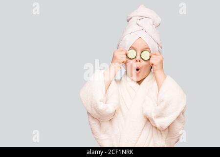 Divertente bambina in camice bianco e asciugamano con maschera per la pelle viso e cetrioli su occhi isolati su sfondo bianco. Trattamenti per la cura della pelle Foto Stock