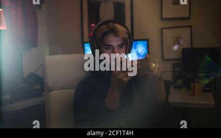 Ragazzo adolescente con cuffie e smartphone in camera oscura Foto Stock