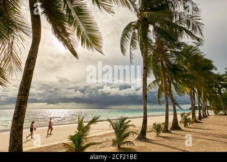 Giovane coppia che cammina lungo la spiaggia bianca pulita con alberi di cocco al tramonto con cielo nuvoloso a Boracay Island, Aklan Province, Visayas, Filippine Foto Stock