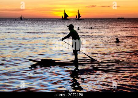 La silhouette di un uomo su una tavola da paddle, la gente che nuota e due barche a vela al tramonto lungo la spiaggia bianca sull'isola di Boracay, Aklan, Filippine. Foto Stock