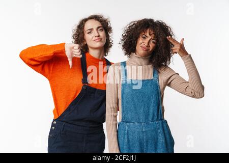 Ritratto di donne multinazionali dispiaciute in tute mostrando il pollice giù e il gesto della pistola isolato su sfondo bianco Foto Stock