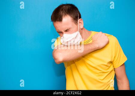 Malato uomo stanco che indossa maschera medica e tosse in gomito isolato su sfondo blu. Coronavirus, Covid-19, persona malata, mal di gola, problema di Foto Stock