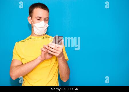 Il giovane indossa maschera medica e t-shirt gialla e l'uso telefono cellulare isolato su sfondo blu Foto Stock