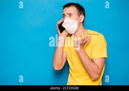 Uomo adulto spaventato nel panico utilizzando smartphone mentre si indossa una maschera chirurgica isolata su sfondo blu. Epidemie, social media, comunicazione e lifesty Foto Stock