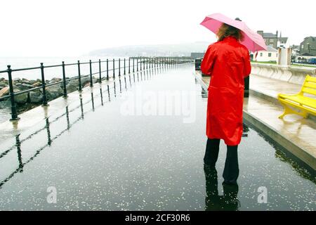 Largs, Ayrshire, Scozia 15 Nov 2003 : Donna in impermeabile rossa comprata da un negozio di beneficenza combatte il vento e la pioggia sulla nuova passeggiata. Seduta su panca gialla, pioggia che corre sopra il suo cappotto Foto Stock