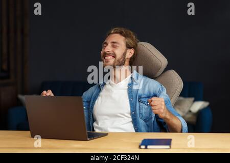 Giovane bell'uomo riccamente dai capelli lunghi che lavora in ufficio con un computer portatile ed esprime positività. Felice uomo d'affari ha ottenuto positivo news.Rewarded, nuovo Foto Stock