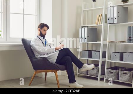 Medico di sesso maschile serio che riempie la scheda del paziente presso il suo ufficio medico. Medico generico che lavora con documenti in ospedale Foto Stock