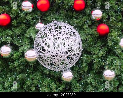 Una grande palla di vimini argentata intrecciata con una ghirlanda pende al centro nei rami di abete con piccolo rosso e palline d'argento intorno Foto Stock