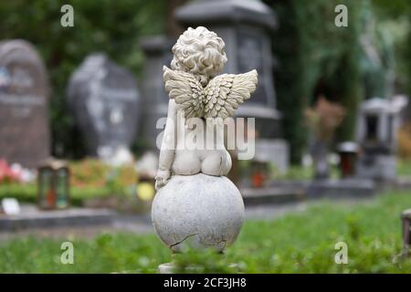 un piccolo angelo è seduto su una palla su un cimitero di fronte a tombe in sfondo sfocato Foto Stock