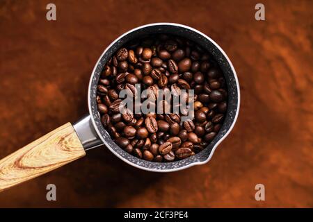 Vista dall'alto della cezve con manico in legno in cui sono tostati chicchi di caffè naturale, e la cezve si trova sulla superficie del caffè macinato. Foto Stock