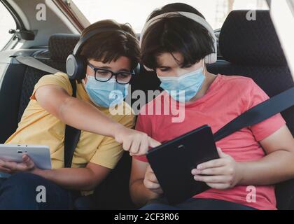 due bambini con maschera medica a cavallo in un'auto da gioco con un tablet Foto Stock