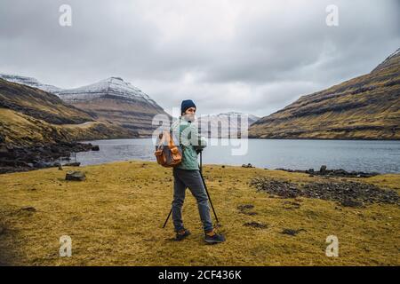 Vista posteriore di uomo bearded in vestiti invernali usando un Fotocamera su un treppiede all'aperto nel paesaggio delle Isole Faroe mentre si guarda la fotocamera Foto Stock