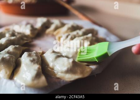 Crop casalinga anonima con spazzola da cucina striscio di torte ripieni di turnover con l'uovo prima di cuocere mentre si prepara la pasta in cucina casalinga Foto Stock