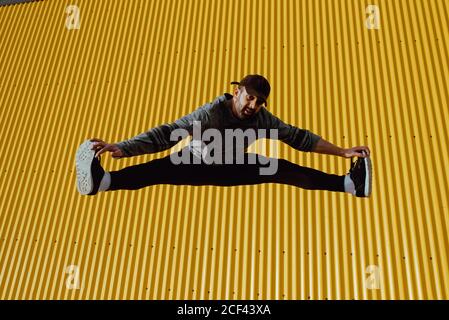 Bel ragazzo bearded in outfit trendy salendo e guardando a macchina fotografica contro il muro giallo di un edificio moderno in pieno sole giorno sulla strada della città Foto Stock