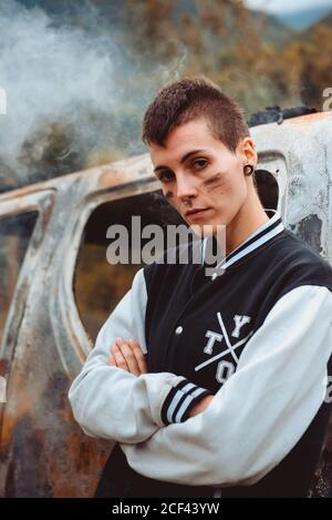 Giovane donna positiva con capelli corti e faccia dipinta che attraversa le braccia e guarda la macchina fotografica mentre si appoggia sulla vecchia auto bruciata in campagna Foto Stock