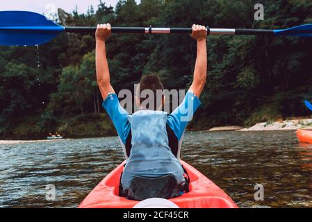 Vista posteriore dello sportivo seduto in canoa rossa e alzando pagaia dopo aver vinto in gara sul declino del fiume Sella in Spagna Foto Stock