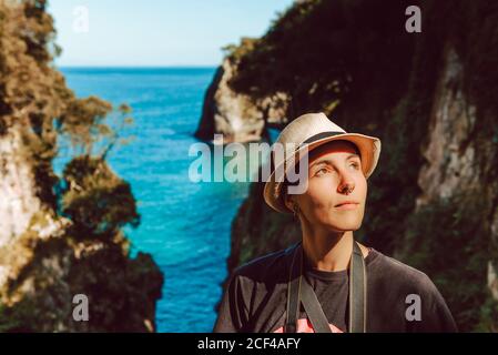 Giovane donna in cappello in piedi con macchina fotografica appesa al collo e godendo di una vista pittoresca del mare e delle rocce a Ribadedeva Asturias Spagna Foto Stock