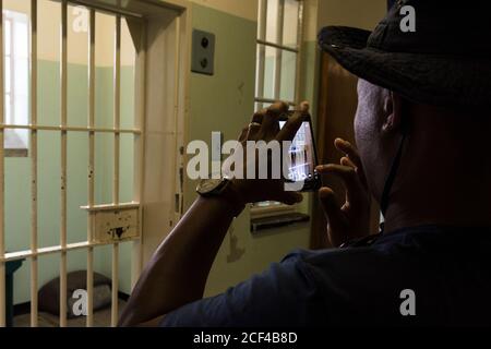 Il turista africano scatta fotografie sul suo smartphone della cella di prigione di Nelson Mandela con i letti disposti. Robben Island al largo di Città del Capo, Sud Africa Foto Stock