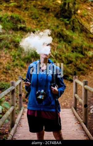 Donna senza volto con macchina fotografica in piedi su ponte di legno dietro fumo spesso mentre fuma smussato durante il campeggio nella foresta Foto Stock