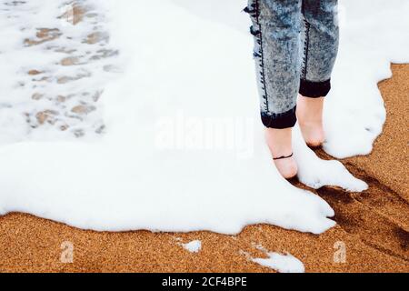 Vista laterale delle gambe di raccolto della femmina a piedi nudi vestita denim in piedi su spiaggia sabbiosa con schiuma di mare Foto Stock