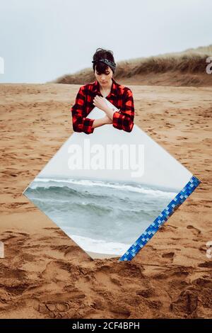 Pensiva femmina unhappy solo giovane in abito casual in piedi spiaggia di sabbia vuota in un giorno cupo e tenendo grande specchio con riflesso di mare tempestoso Foto Stock