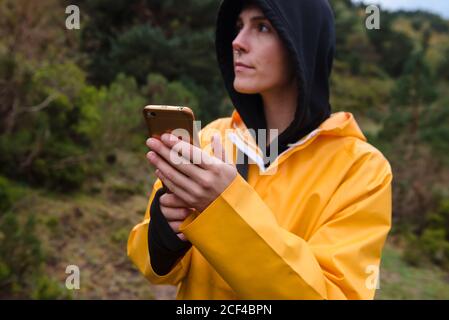 Giovane donna premurosa con felpa con cappuccio scura e impermeabile giallo con smartphone che guarda lontano nella foresta Foto Stock