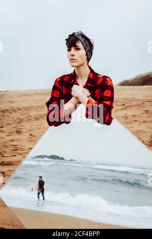 Premurosa giovane donna triste in camicia casual a scacchi in piedi spiaggia di sabbia e grande specchio con riflesso di tempesta mare e uomo che cammina Foto Stock