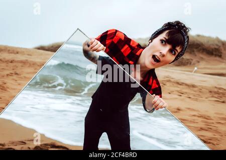 Sorpresa scioccata giovane donna in abito casual in piedi su sabbia spiaggia e tenendo grande specchio con riflesso di maschio raccolto contro il mare tempestoso Foto Stock