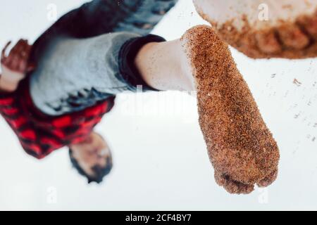 Da sotto vista capovolta di giovane hipster femmina in camicia casual a scacchi e jeans con sabbia bagnata sui piedi si riflette sulla finestra che si erge sulla spiaggia Foto Stock