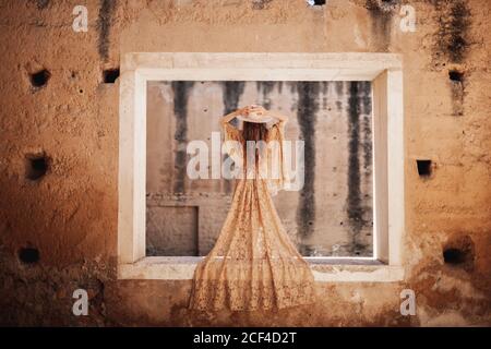 Vista posteriore della splendida donna in pizzo beige vestito in piedi sulla parete di un edificio deserto e tenendo il cappello Foto Stock
